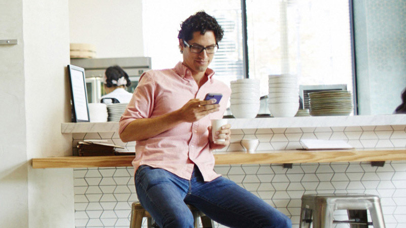 Man sitting at counter looking at mobile phone and holding a cup of coffee.